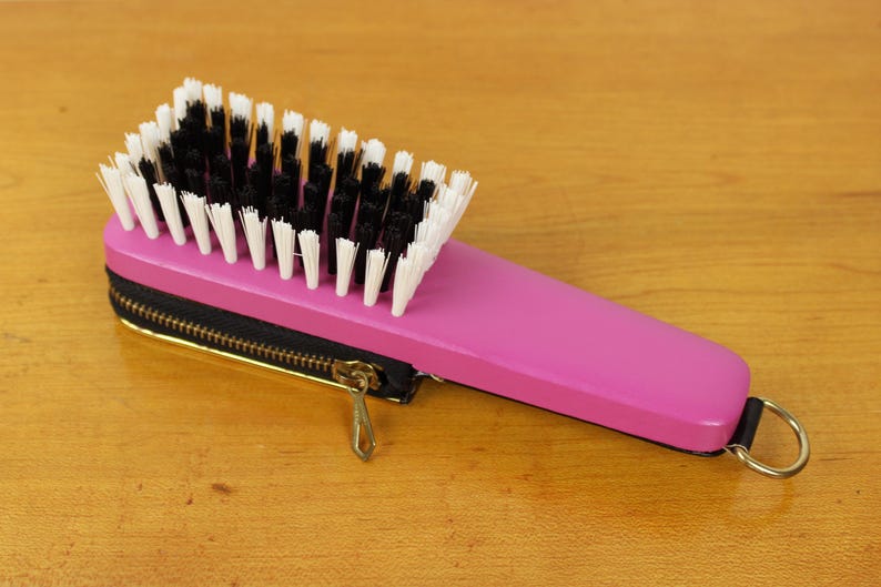 May include: A pink hairbrush with a black zipper case and a gold-colored zipper pull. The brush has black and white bristles and a handle with a gold ring.