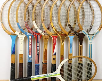 May include: A collection of ten vintage wooden tennis rackets arranged in a row, with nine standing upright and one positioned horizontally in front. The rackets feature various Wilson branding, colorful painted accents, and black grip tape.