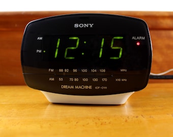 May include: A black Sony Dream Machine ICF-C111 alarm clock with a white base. The digital display shows the time 12:15 in bright green numbers. The front panel features AM and FM radio frequency scales and a small red alarm indicator light.
