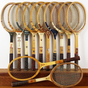 May include: A collection of eleven vintage wooden tennis rackets arranged with nine standing upright in a row and two positioned horizontally in the foreground. Each racket features a wooden frame, nylon stringing, and a wrapped handle grip.