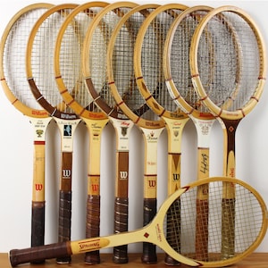 May include: A collection of eight vintage wooden tennis rackets arranged in a row with one positioned horizontally in the foreground. The rackets feature light and dark wood frames, nylon string patterns, and various brand logos including Wilson and Spalding.