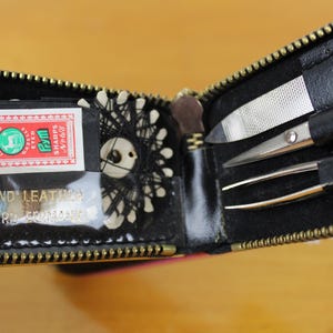 May include: Open black leatherette sewing kit with a gold zipper. The kit contains a small box of sewing needles, thread, scissors, tweezers, and a nail file. The box of needles has the text "SHARPS No. 8".
