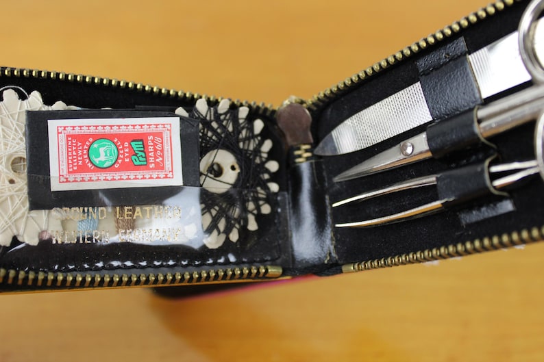 May include: Open black leatherette sewing kit with a gold zipper. The kit contains a small box of sewing needles, thread, scissors, tweezers, and a nail file. The box of needles has the text "SHARPS No. 8".