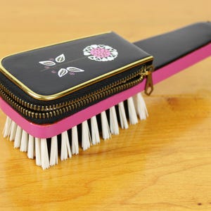 May include: A vintage black and pink brush with a zippered top featuring a floral design. The brush has a gold zipper and trim, with white bristles. The handle is black and pink, and the brush is set on a wooden surface.