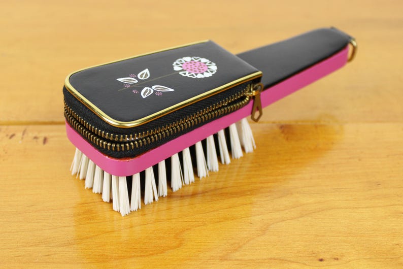 May include: A vintage black and pink brush with a zippered top featuring a floral design. The brush has a gold zipper and trim, with white bristles. The handle is black and pink, and the brush is set on a wooden surface.