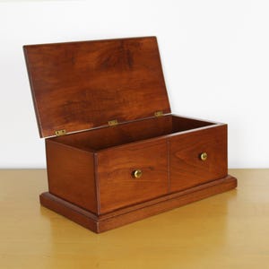 May include: A wooden box with a hinged lid, open to reveal an empty interior. The box is a rich, warm brown color with two small drawers on the front, each with a round, brass-colored knob. The box sits on a rectangular base.