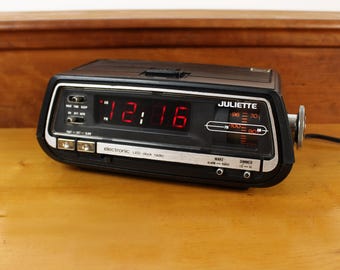 May include: A black Juliette electronic LED clock radio featuring a digital display showing 12:16 in red, an analog AM/FM radio tuner dial, and various control switches for alarm, sleep, and wake functions on the front panel.
