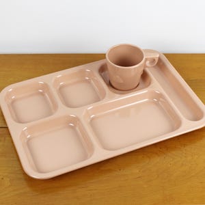 May include: A peach-colored divided tray with five compartments and a matching mug with a handle. The tray is made of a smooth, glossy material, perfect for serving meals. The mug sits in a designated circular space on the tray.