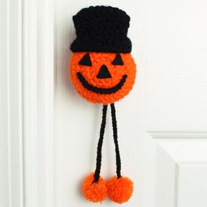 May include: A crocheted jack-o'-lantern decoration featuring a bright orange pumpkin head with a black felt face, topped with a black crocheted hat. Two long black yarn legs extend downward, each ending in a fluffy orange yarn pom-pom foot.