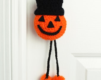 Puede incluir: Una decoración de calabaza tejida a ganchillo con una cabeza naranja brillante, un rostro de fieltro negro y un sombrero de copa negro. Dos largas piernas de hilo negro cuelgan hacia abajo, terminando en pompones de lana naranja.