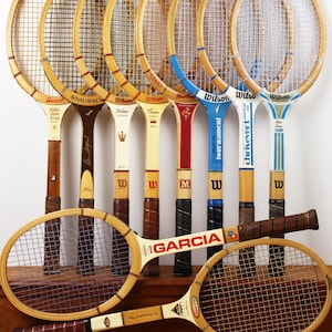May include: A collection of vintage wooden tennis rackets arranged in a row with two rackets positioned horizontally in the foreground. The rackets feature various brand names including Wilson, Bancroft, Snauwaert, and Garcia, with colorful accents.