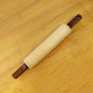 May include: A wooden rolling pin with a light-colored, cylindrical rolling surface and dark brown handles. The handles are attached to the rolling surface and have a hole at the end for hanging. The rolling pin is made of wood and is designed for baking.