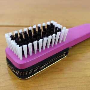 May include: A pink brush with black and white bristles, attached to a black zippered case with a gold zipper. The brush has a small gold ring on the handle. The item is on a wooden surface.