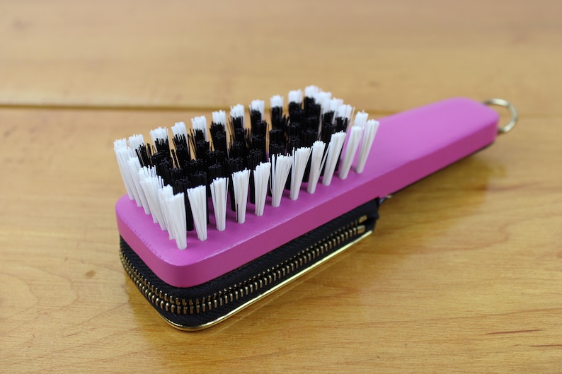 May include: A pink brush with black and white bristles, attached to a black zippered case with a gold zipper. The brush has a small gold ring on the handle. The item is on a wooden surface.