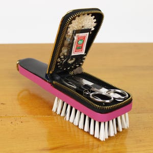 May include: A pink and black manicure set with a brush base. The set includes scissors, nail files, and other grooming tools. The case is black with a gold zipper and a pink base with white bristles.