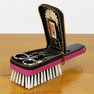 May include: A vintage travel sewing kit with a pink base and a black zippered case. The case contains small scissors and other sewing tools. The kit is attached to a brush with white bristles.