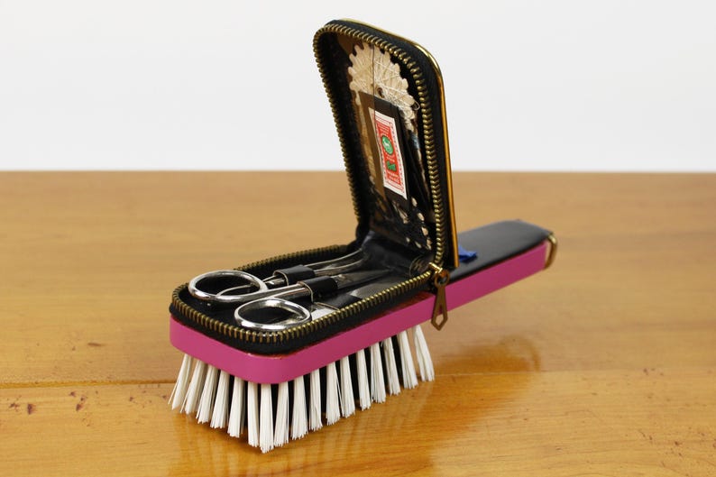 May include: A vintage travel sewing kit with a pink base and a black zippered case. The case contains small scissors and other sewing tools. The kit is attached to a brush with white bristles.