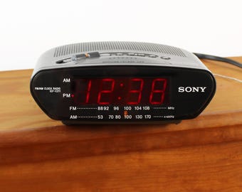 May include: A black Sony FM/AM clock radio model ICF-C211 with a digital display showing the time 12:38 in bright red LED numbers. The front panel features AM and PM indicators, radio frequency scales for FM and AM bands, and a power cord.