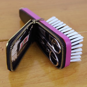 May include: A black zippered case with gold trim, open to reveal small scissors and other tools, is positioned next to a pink and white nail brush. The case and brush are on a light brown wooden surface.