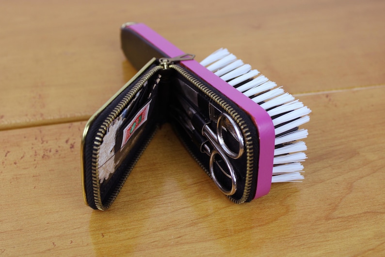 May include: A black zippered case with gold trim, open to reveal small scissors and other tools, is positioned next to a pink and white nail brush. The case and brush are on a light brown wooden surface.