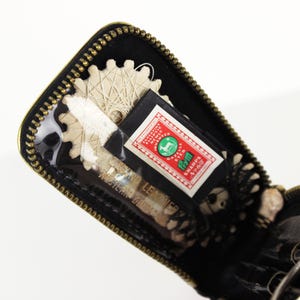 May include: Open black zippered case with gold trim, containing sewing tools. Visible are scissors, a spool of thread, and a label with text and a green emblem. The case has a clear plastic window.