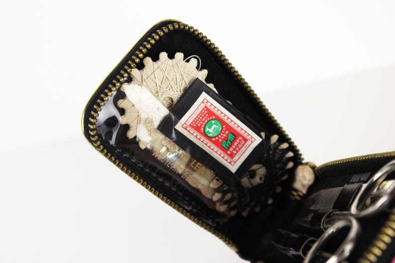 May include: Open black zippered case with gold trim, containing sewing tools. Visible are scissors, a spool of thread, and a label with text and a green emblem. The case has a clear plastic window.