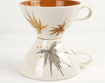 May include: Two stacked ceramic cups with a wide, flared rim and a small handle. The top cup has a cream exterior with brown leaf illustrations and a solid orange interior. The bottom cup features a cream exterior with a grey leaf illustration.
