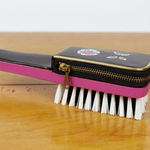 May include: A vintage hairbrush with a black and pink handle and a zippered compartment. The brush features a black top with a floral design and gold trim, and white bristles.