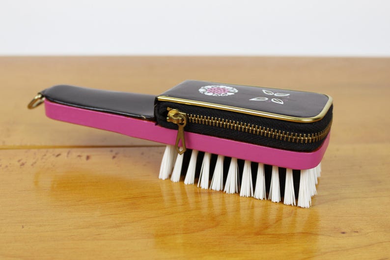 May include: A vintage hairbrush with a black and pink handle and a zippered compartment. The brush features a black top with a floral design and gold trim, and white bristles.