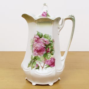 May include: A white porcelain pitcher with a lid, handle, and spout. The pitcher is decorated with a large pink rose floral design and green leaves. The handle and rim have a green and gold trim. The pitcher sits on a wooden surface.