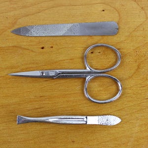 May include: A manicure set with a nail file, small scissors, and a cuticle pusher. The tools are made of silver-colored metal and are arranged on a wooden surface.