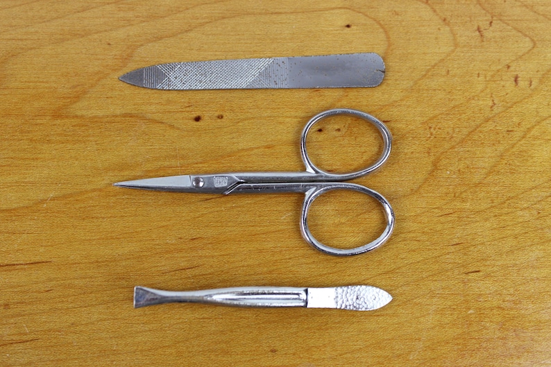 May include: A manicure set with a nail file, small scissors, and a cuticle pusher. The tools are made of silver-colored metal and are arranged on a wooden surface.
