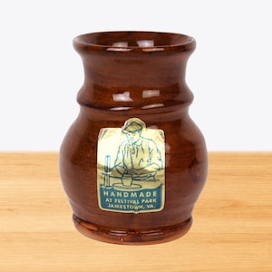 May include: A handmade brown ceramic vase with a glossy finish. The vase has a unique shape with a wider base and a narrower neck. A sticker on the front features an illustration of a craftsman and the text "HANDMADE AT FESTIVAL PARK JAMESTOWN, VA."