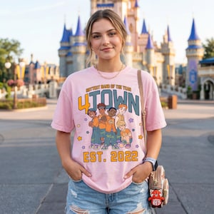 Può includere: T-shirt rosa chiaro con la scritta "Hottest Band of the Year 4*TOWN EST. 2022" e una grafica di cinque personaggi dei cartoni animati. La maglietta è indossata da una persona davanti a un castello.