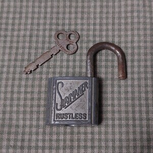 Puede incluir: Un candado vintage Slaymaker Rustless con una llave. El candado es plateado con letras negras y la llave está oxidada.