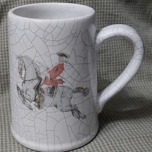 May include: White ceramic mug with a crackled glaze. The mug features a colorful illustration of a person riding a horse.