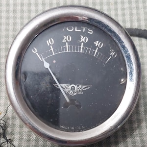 May include: A vintage silver-colored analog voltmeter with a black face. The gauge reads "VOLTS" and has markings from 0 to 50. The needle is pointing to 10. The gauge has a silver winged logo with the text "JEWELL" below it. The text "MADE IN U.S.A." is visible at the bottom of the gauge.