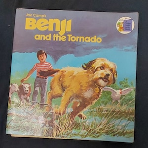 May include: A colorful book cover featuring a brown dog running through a field with a boy and two cows. The book title is "Benji and the Tornado" by Joe Camp.
