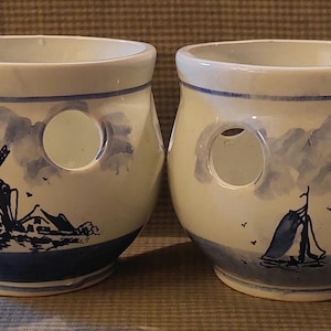 May include: Two ceramic pots with a blue and white Dutch design. Each pot has three circular openings and is decorated with a windmill, houses, and a sailboat scene. The pots have a blue base and rim.