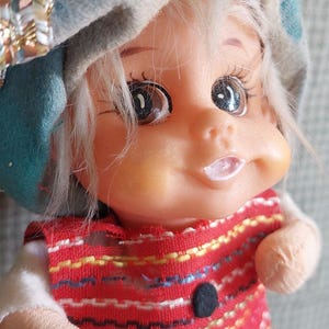 May include: A vintage doll with a painted face, white hair, and a red vest with black buttons. The doll wears a blue and gray hat with a decorative trim. The doll's eyes are large and expressive.