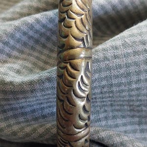 May include: A cylindrical, antique-looking metal object with an intricate, repeating pattern of curved lines and shapes. The object has a tarnished, bronze-like appearance and is set against a textured, gray-green fabric.
