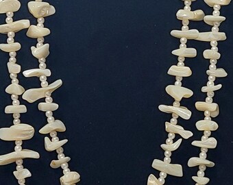 May include: A long, multi-strand necklace made of white shell beads and small white pearls. The necklace is layered and hangs down the front of the neck.