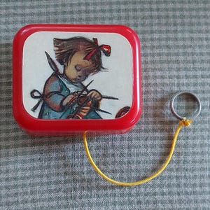 May include: A vintage red and white music box featuring an illustration of a young girl knitting. The box has a silver ring attached with a yellow string. The background is a gray and white checkered pattern.