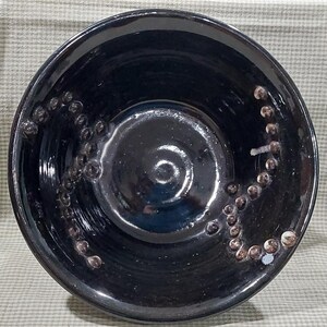 May include: A handmade, black ceramic bowl with a glossy finish. The bowl features a spiral design in the center and a decorative pattern of small, raised, brown-colored dots around the rim. The bowl is round and has a deep interior.