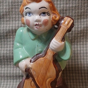 May include: A ceramic figurine of a person playing a cello. The figurine is painted in shades of green, brown, and white. The person has red hair and is wearing a green shirt. The cello is brown with a light brown bow.