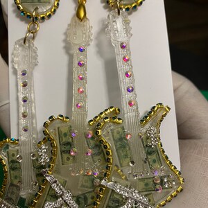 May include: A pair of clear guitar-shaped earrings with a gold trim and rhinestones. The earrings are filled with images of US dollar bills and feature a silver gun charm.