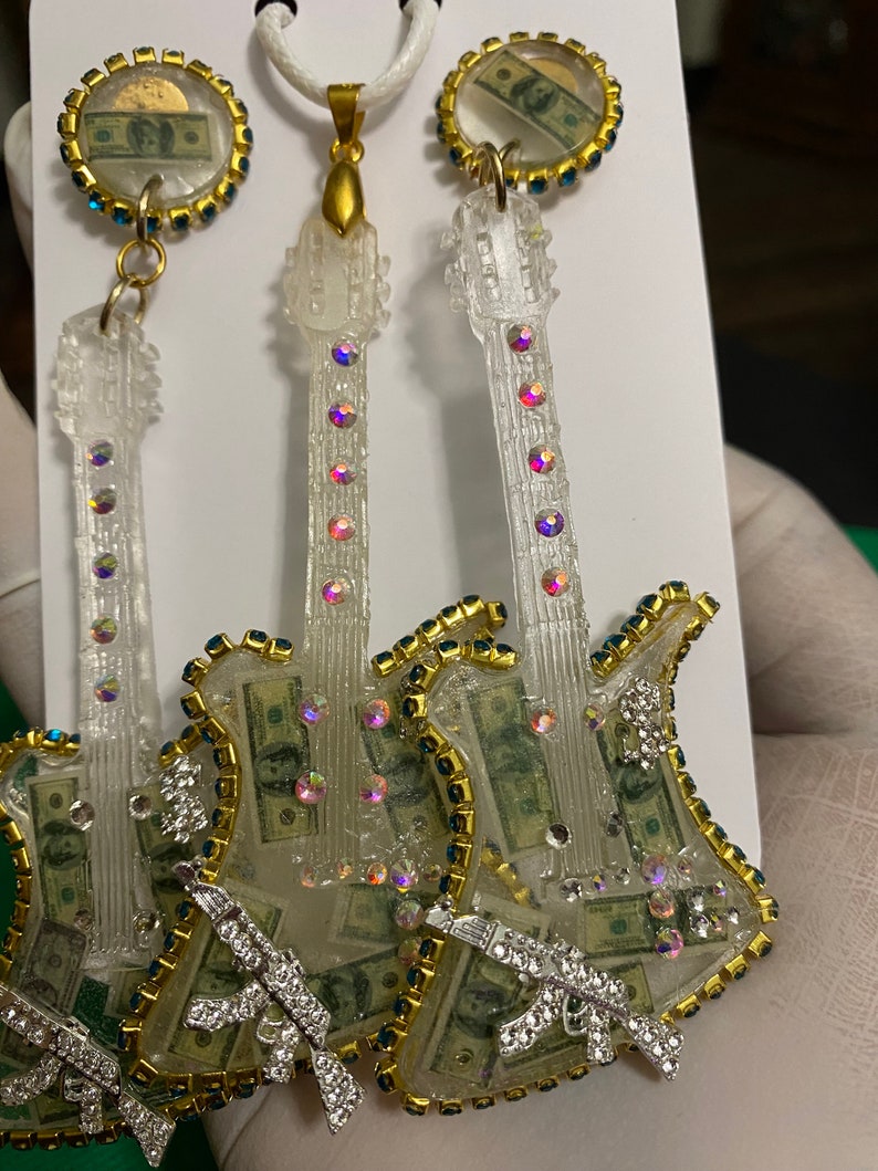 May include: A pair of clear guitar-shaped earrings with a gold trim and rhinestones. The earrings are filled with images of US dollar bills and feature a silver gun charm.