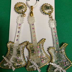 May include: A set of three clear guitar-shaped earrings with gold accents and rhinestones. Each earring features a dollar bill inside and a silver gun charm. The set also includes a matching pendant.