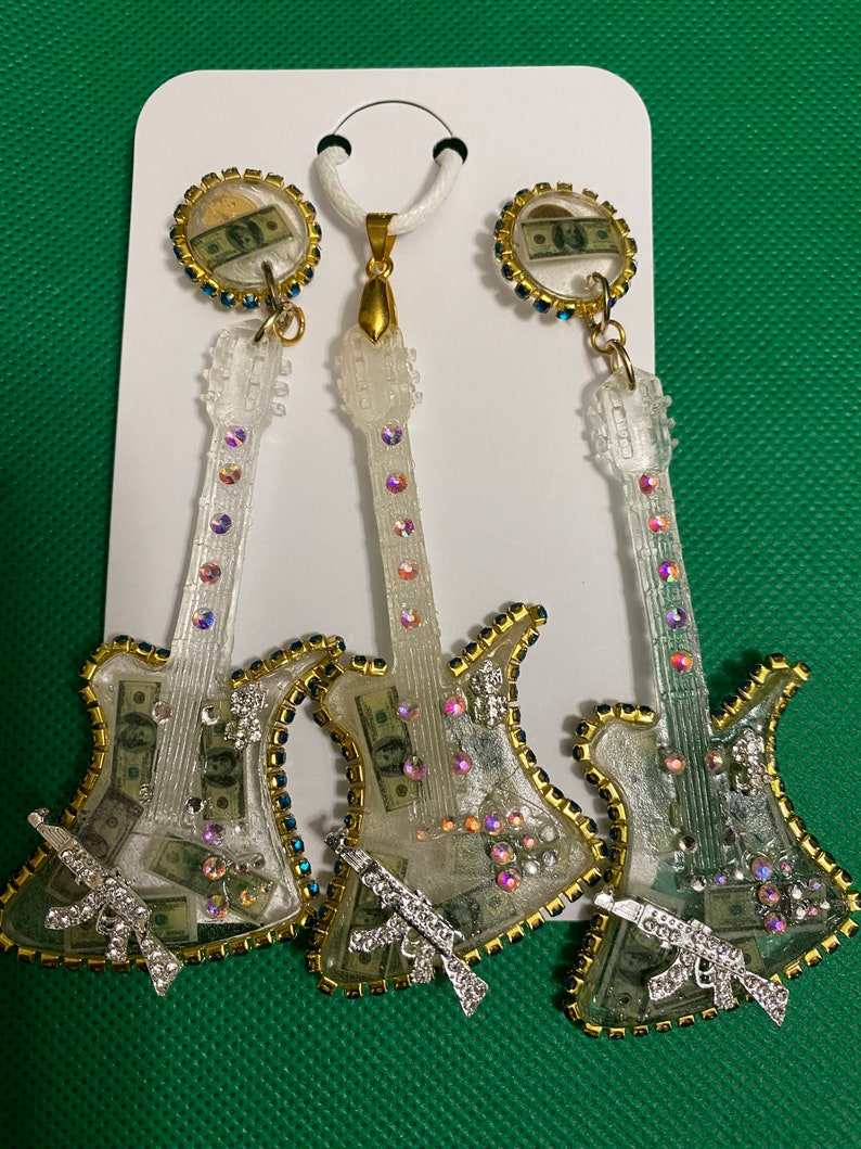 May include: A set of three clear guitar-shaped earrings with gold accents and rhinestones. Each earring features a dollar bill inside and a silver gun charm. The set also includes a matching pendant.