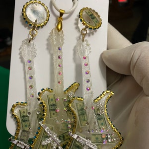 May include: Three clear resin guitar earrings with gold trim and rhinestones. The guitars are filled with images of US dollar bills and have a silver gun charm on each. The earrings are hanging from gold hoops with a clear resin circle with a US dollar bill image.